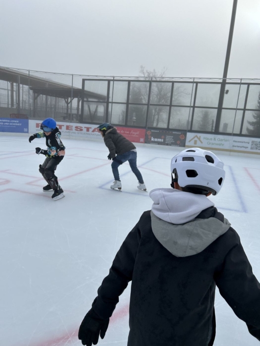 Zwei Kinder spielen Fangen auf dem Eisfeld, ein drittes schaut zu.